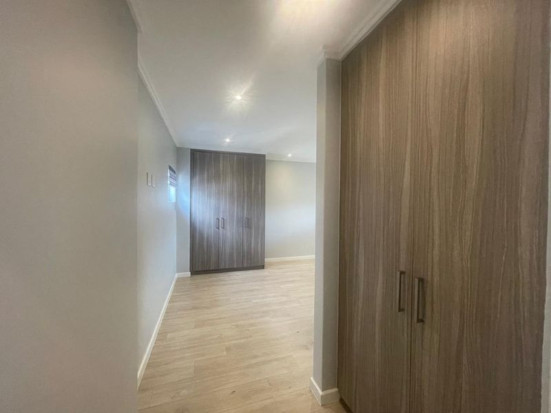 A bright, modern bedroom featuring neutral walls, light laminate flooring, and extensive built-in cupboard storage with a contemporary wood-grain finish.