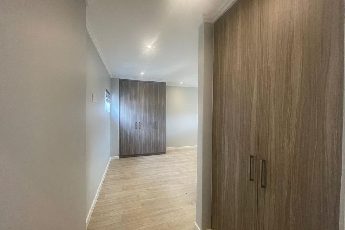 A bright, modern bedroom featuring neutral walls, light laminate flooring, and extensive built-in cupboard storage with a contemporary wood-grain finish.