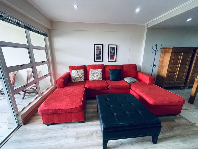 A bright living room featuring modern laminate flooring, recessed downlighting, and large windows that provide ample natural light and access to the balcony.