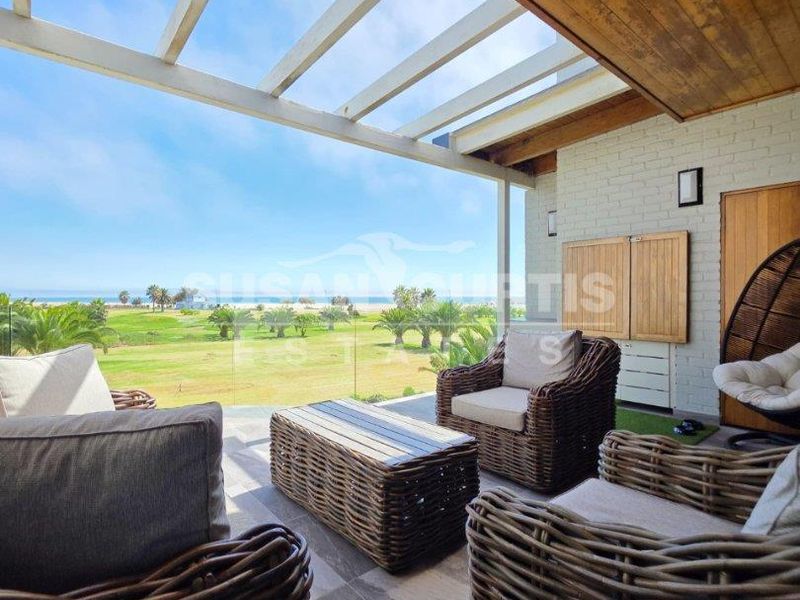 A covered patio featuring a timber-clad ceiling, pergola, and glass balustrade, offering scenic views of the surrounding landscape and coastline.