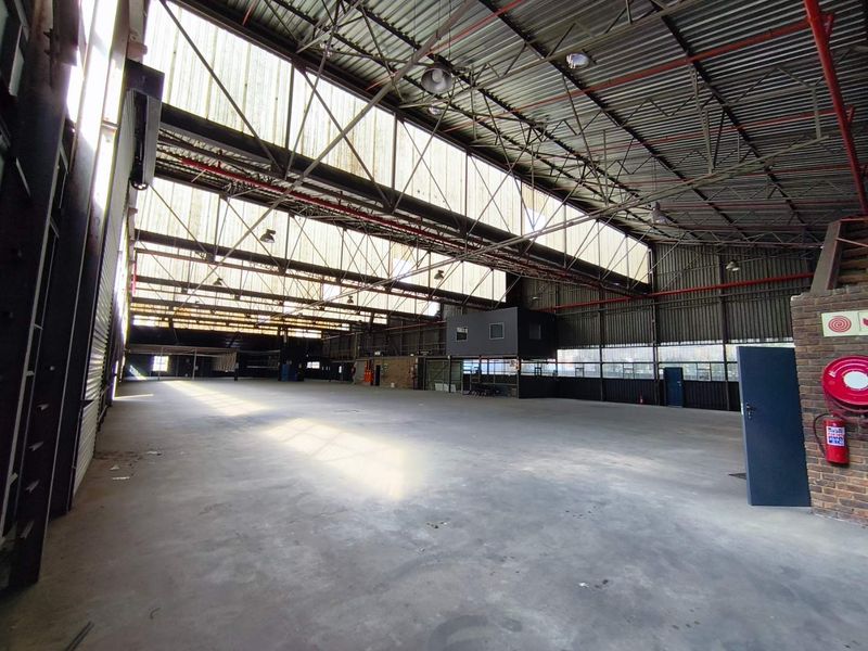 Spacious industrial warehouse featuring high ceilings with exposed steel trusses, translucent roof panels for natural light, concrete flooring, and an integrated mezzanine office area.