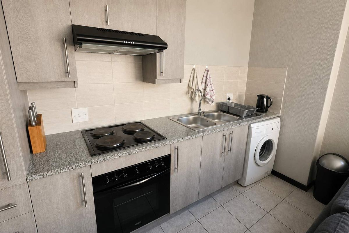Compact modern kitchen featuring light wood-grain cabinetry, granite countertops, built-in electric oven and hob, extractor fan, and integrated laundry plumbing.