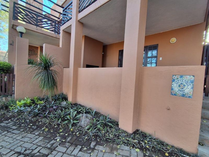 A covered ground-floor patio area featuring a built-in braai, adjacent to a paved driveway and landscaped garden bed.