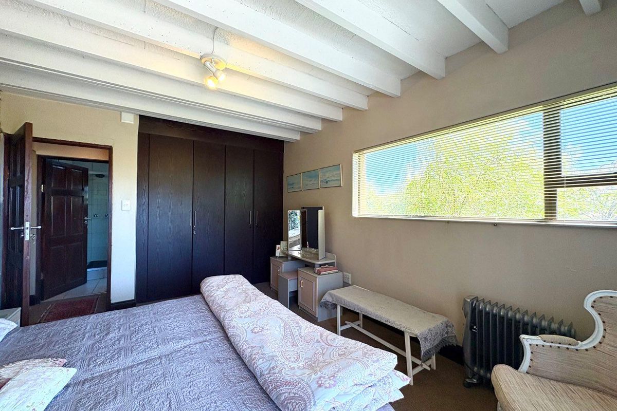 A bedroom featuring exposed white timber ceiling beams, built-in dark wood cabinetry, and a horizontal window with integrated blinds.