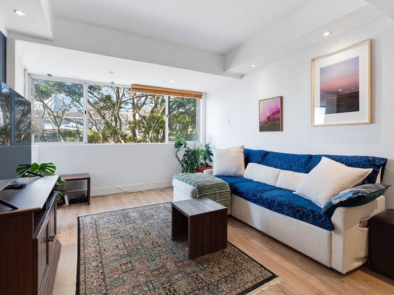 A bright and well-maintained living room featuring laminate flooring, recessed downlighting, and a large window providing natural light and a pleasant leafy outlook.