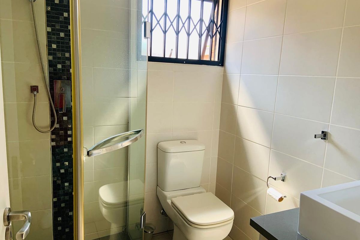 A functional bathroom featuring a glass-enclosed shower, a modern white toilet, and a vanity basin with tiled walls and security bars on the window.