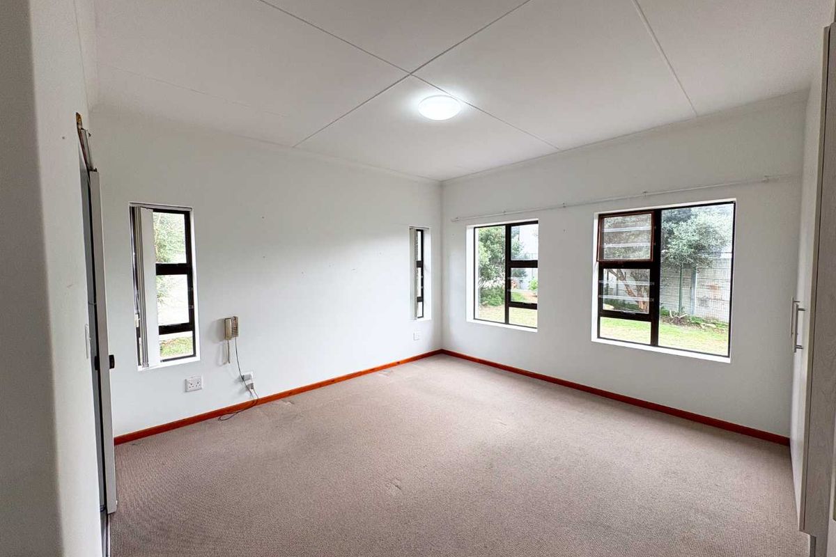 A bright, empty bedroom featuring neutral carpeting, built-in cupboards, and multiple windows providing natural light and garden views.