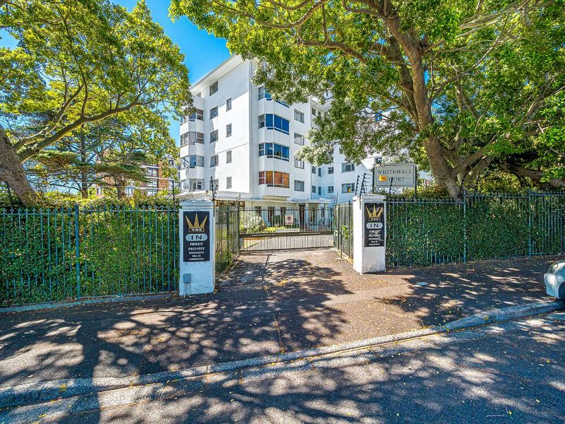 Street-level view of the Whitehall Court apartment complex entrance, featuring secure gated access and mature trees surrounding the building facade.