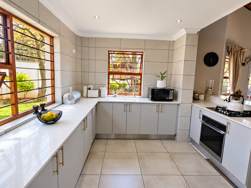 Modern kitchen featuring light grey cabinetry, quartz countertops, built-in oven, gas hob, and large windows with security bars overlooking the garden.