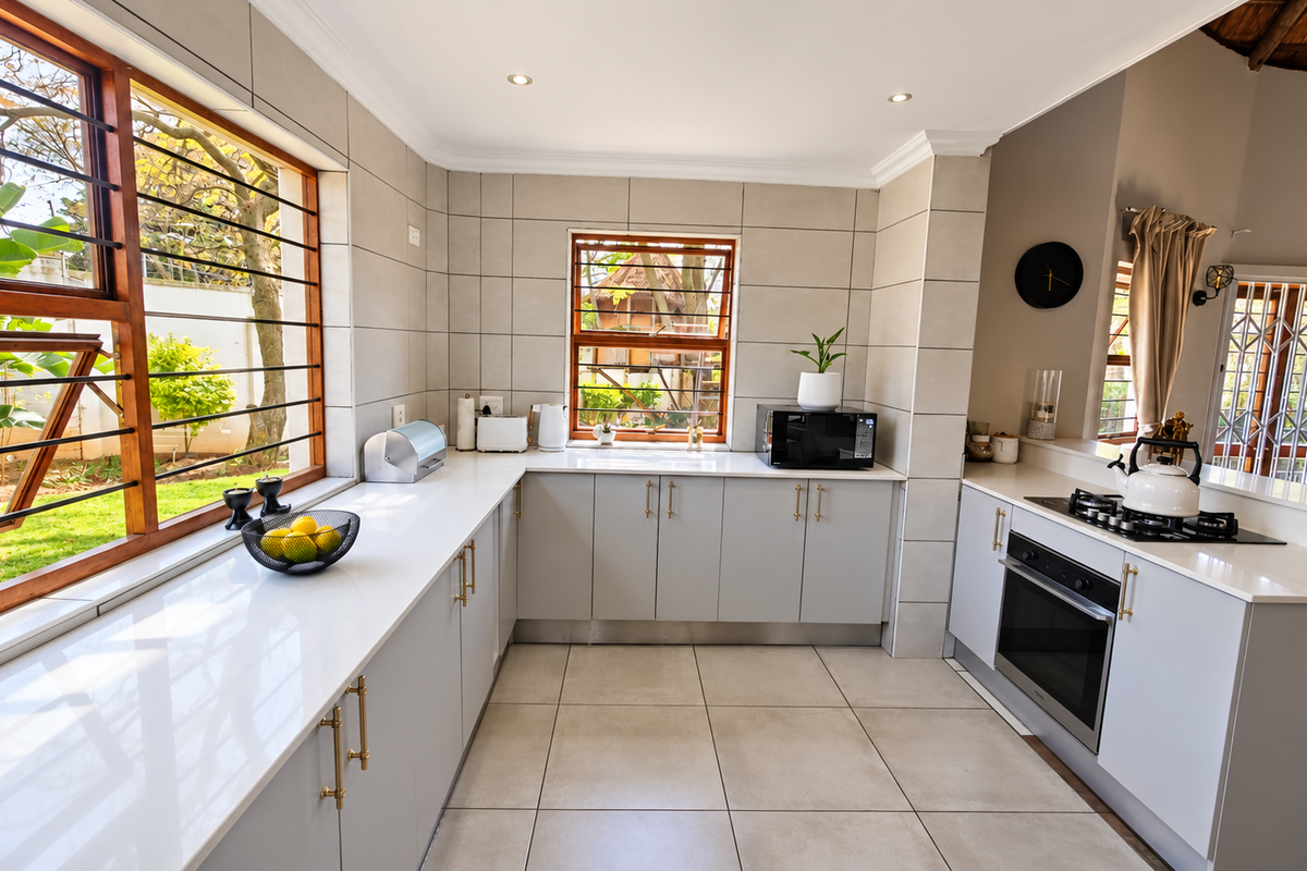 Modern kitchen featuring light grey cabinetry, quartz countertops, built-in oven, gas hob, and large windows with security bars overlooking the garden.