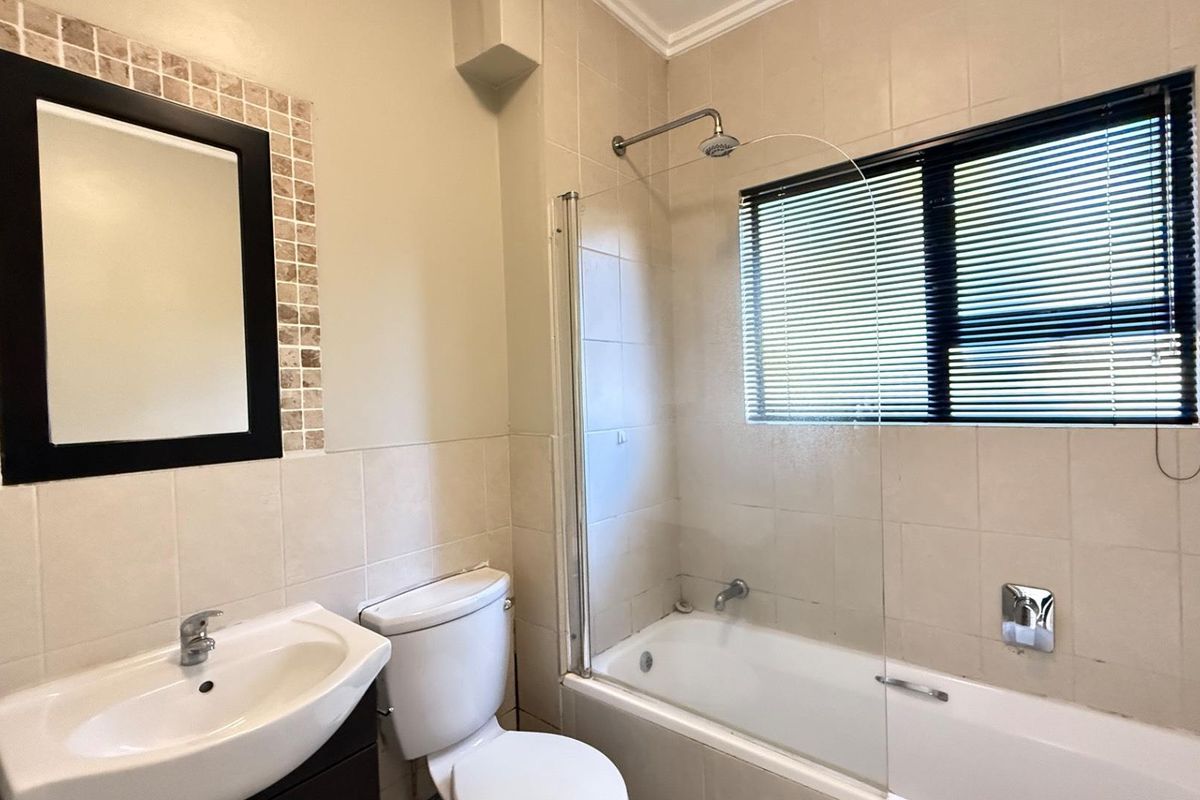 A clean, functional bathroom featuring a bathtub with an integrated shower, glass screen, pedestal basin, and neutral wall tiling.