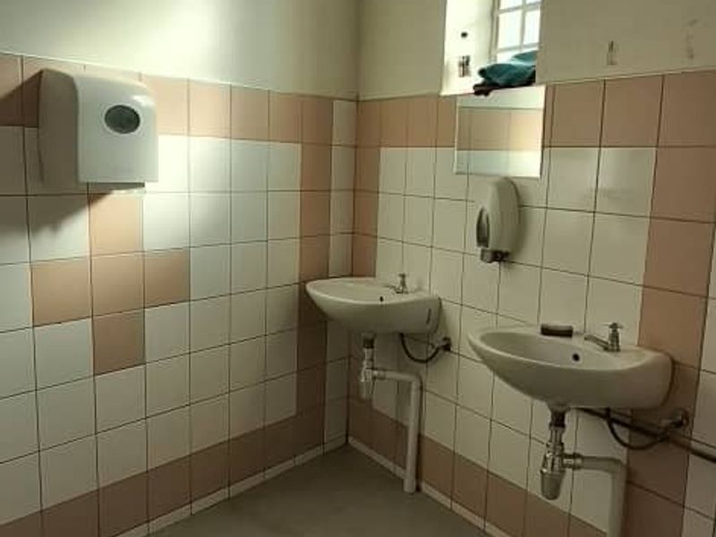 A functional commercial-style bathroom featuring dual wall-mounted washbasins, tiled walls, and tiled flooring, requiring cosmetic updates to improve its dated appearance.