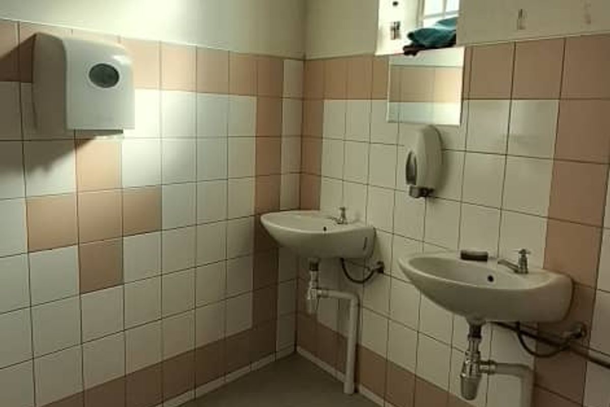 A functional commercial-style bathroom featuring dual wall-mounted washbasins, tiled walls, and tiled flooring, requiring cosmetic updates to improve its dated appearance.