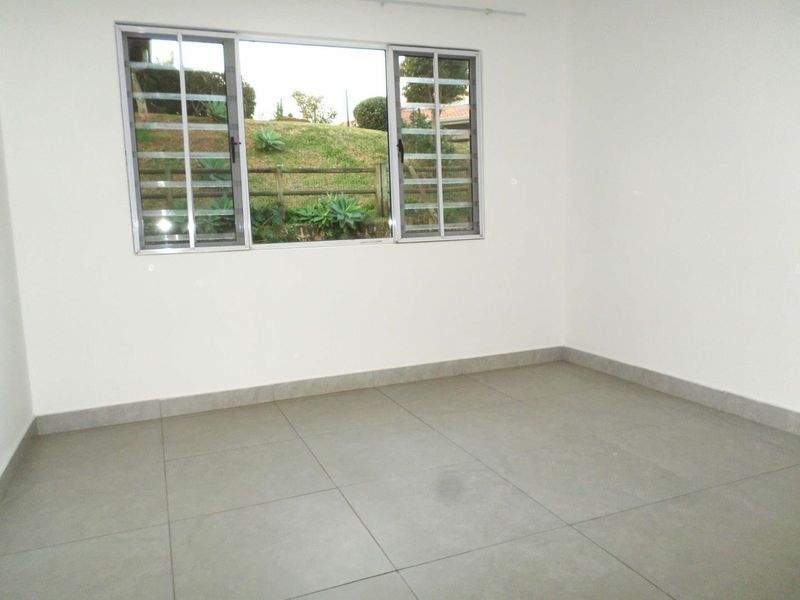 A bright, empty bedroom featuring neutral tiled flooring, clean white walls, and aluminium-framed windows with security bars overlooking the garden.