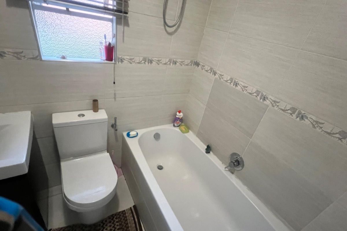 A functional bathroom featuring a bathtub, wall-mounted shower head, toilet, and vanity basin with neutral wall tiling and a frosted glass window.