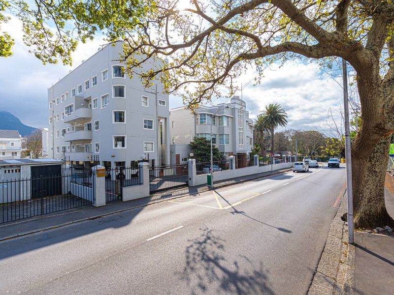Street-level view of the apartment building facade, featuring multi-story architecture, secure perimeter fencing, and gated access.
