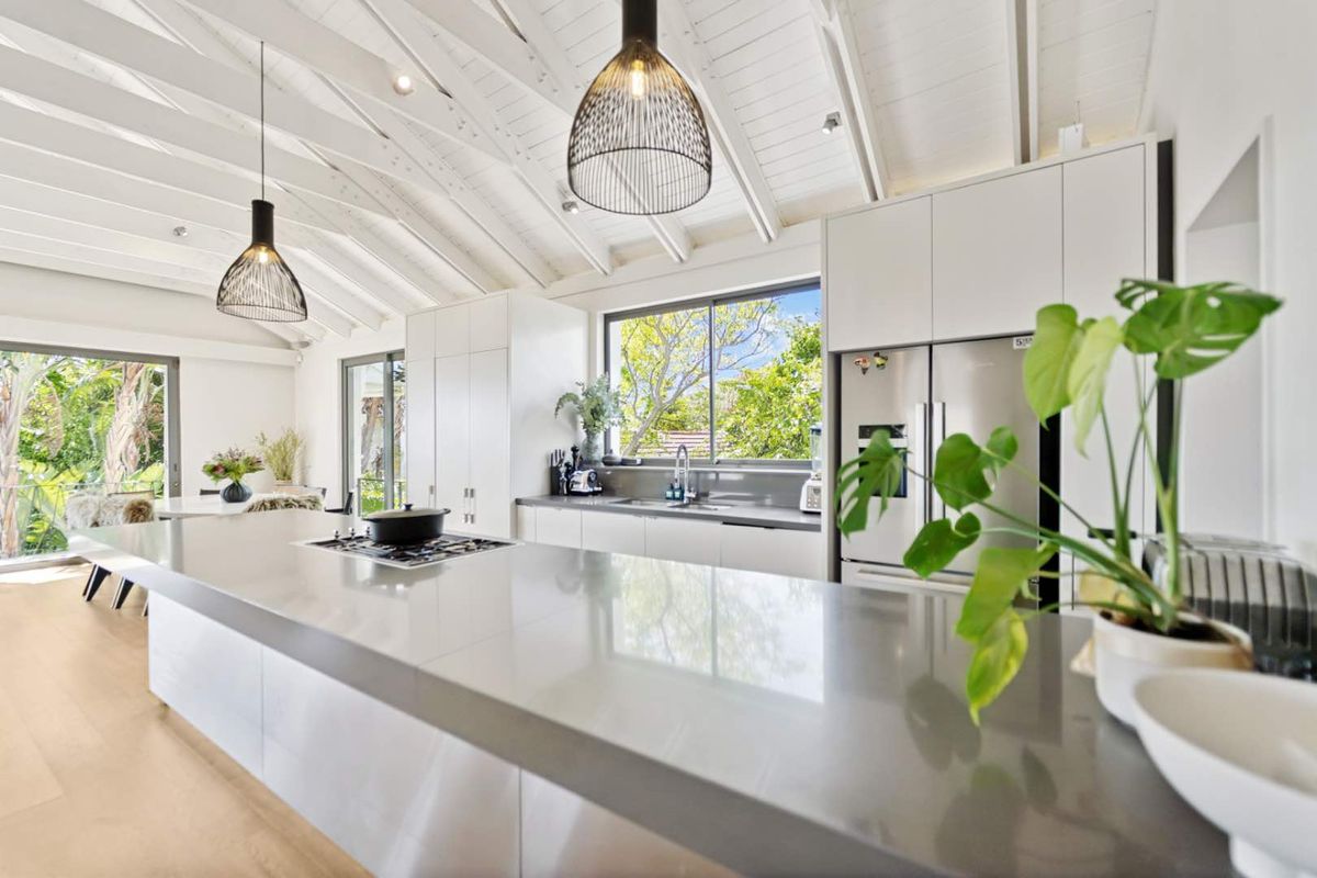 Modern kitchen featuring a large stone island, gas cooktop, integrated cabinetry, and a striking vaulted ceiling with exposed beams, opening to a lush garden view.