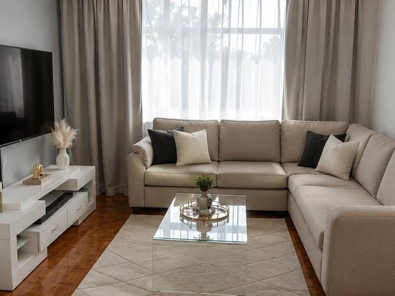 A well-maintained living room featuring warm parquet flooring, a large window with neutral curtains, and a clean, modern aesthetic.