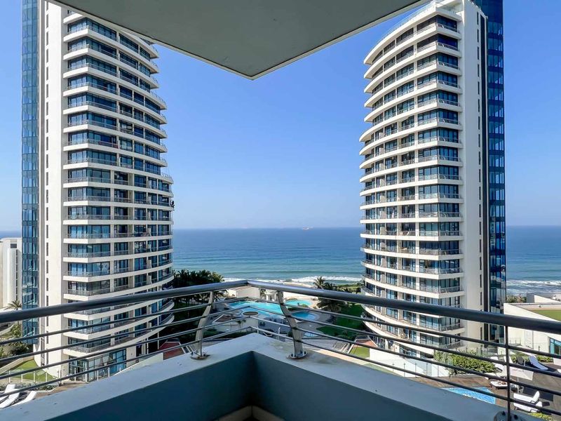 A balcony view overlooking the ocean and surrounding high-rise apartment complex, featuring a metal railing and clear coastal outlook.