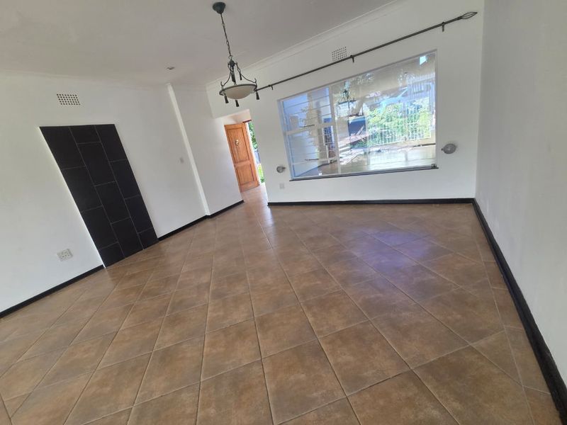 A spacious, empty living room featuring neutral tiled flooring, a large window for natural light, and a decorative black tiled feature wall.