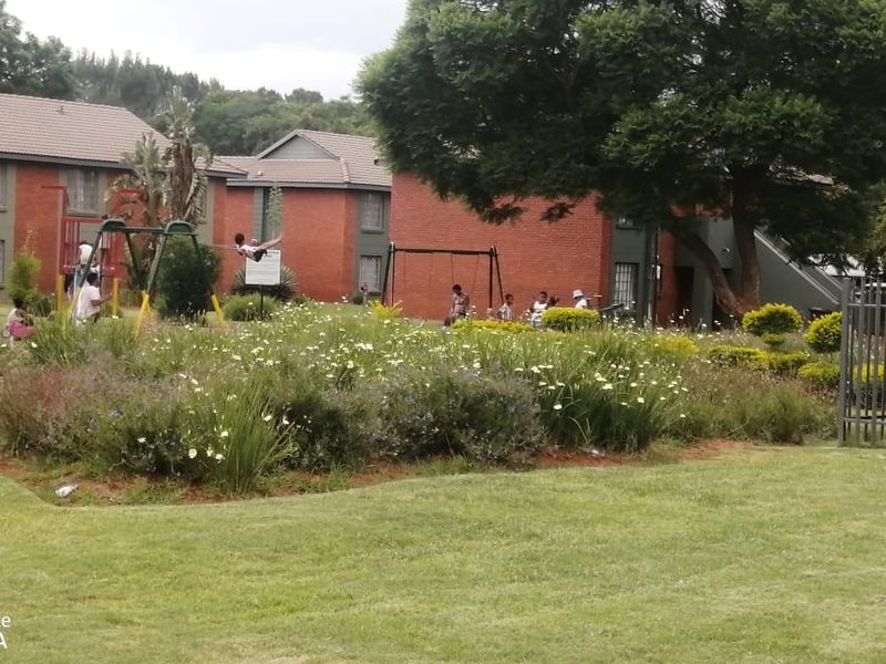 A communal garden area featuring landscaped lawns, established trees, and playground equipment situated between brick apartment buildings.