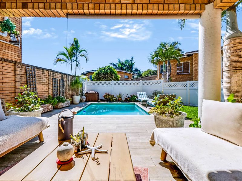 A well-maintained outdoor entertainment area featuring a swimming pool, paved patio, and covered seating space enclosed by brick and vinyl fencing.