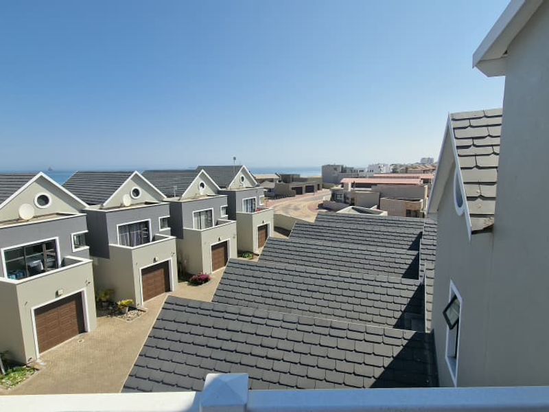 Elevated view overlooking a residential townhouse complex with tiled roofs, paved driveways, and a distant ocean horizon.