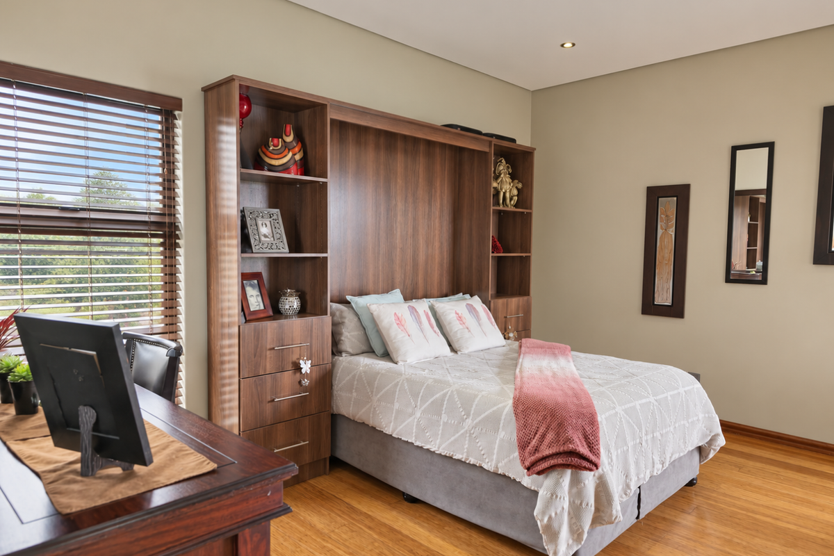 A well-maintained bedroom featuring warm timber flooring, recessed lighting, and a custom built-in headboard shelving unit with integrated storage.