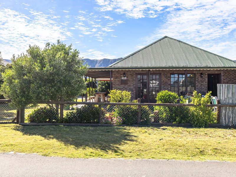 A charming single-story face brick house featuring a corrugated metal roof, timber-framed windows, and a covered patio area, set behind a rustic timber fence.