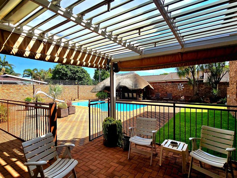 Covered patio area featuring a louvered roof and retractable awning, overlooking a private swimming pool, garden, and a traditional thatched lapa.