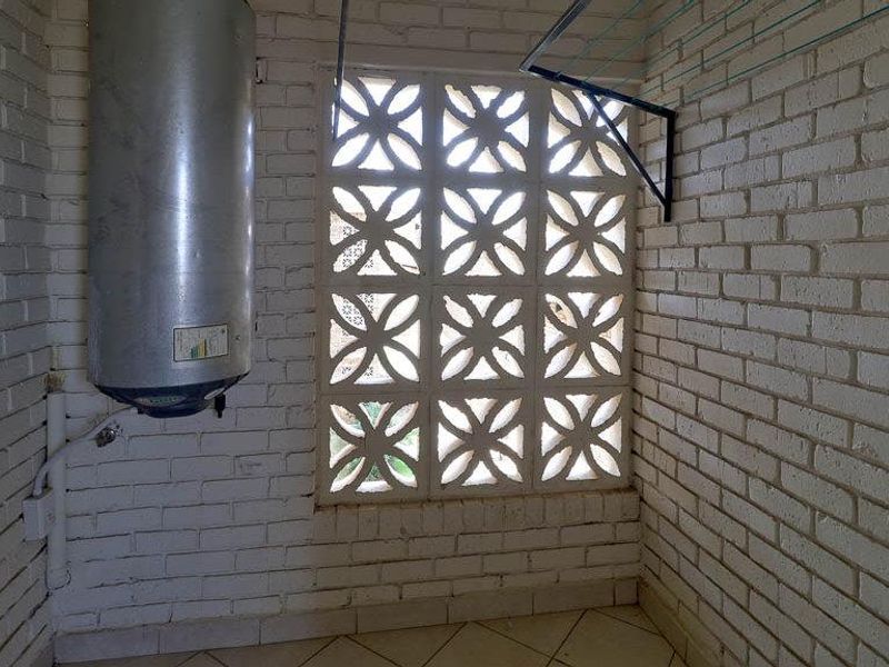A functional laundry area featuring exposed brick walls, a wall-mounted water heater, tiled flooring, and a decorative breeze block window for ventilation.