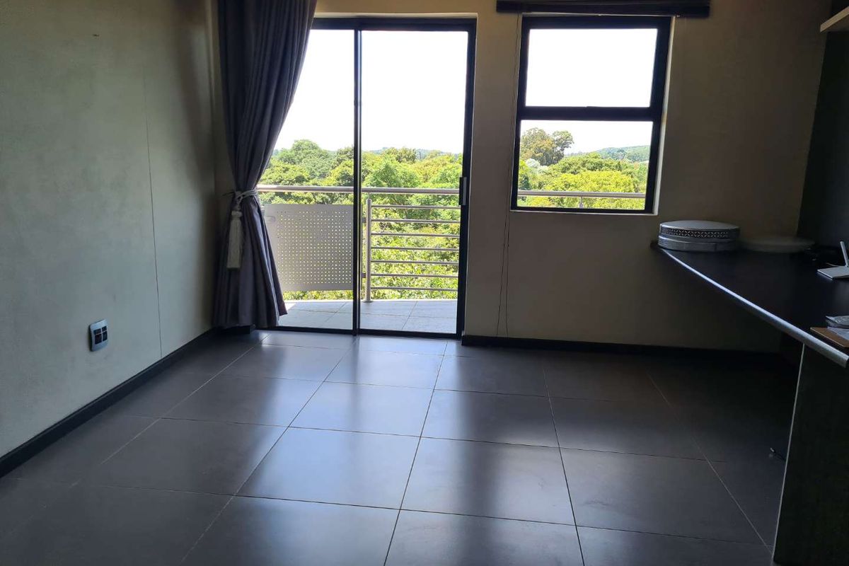 An empty living area featuring large format floor tiles, a built-in desk, and sliding glass doors opening onto a balcony with scenic views.