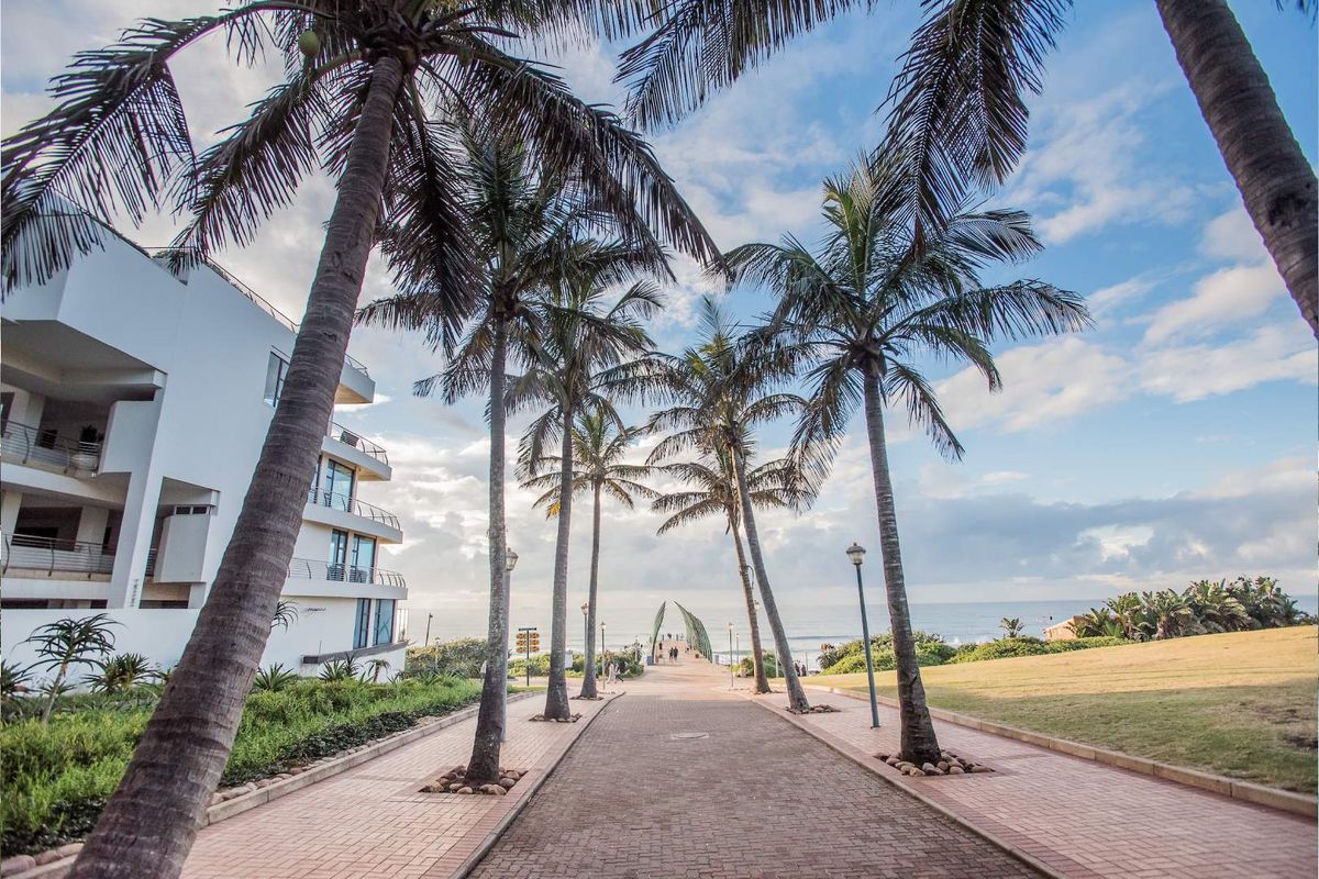 A scenic paved walkway lined with palm trees leading towards the ocean, adjacent to a modern apartment building with balconies.