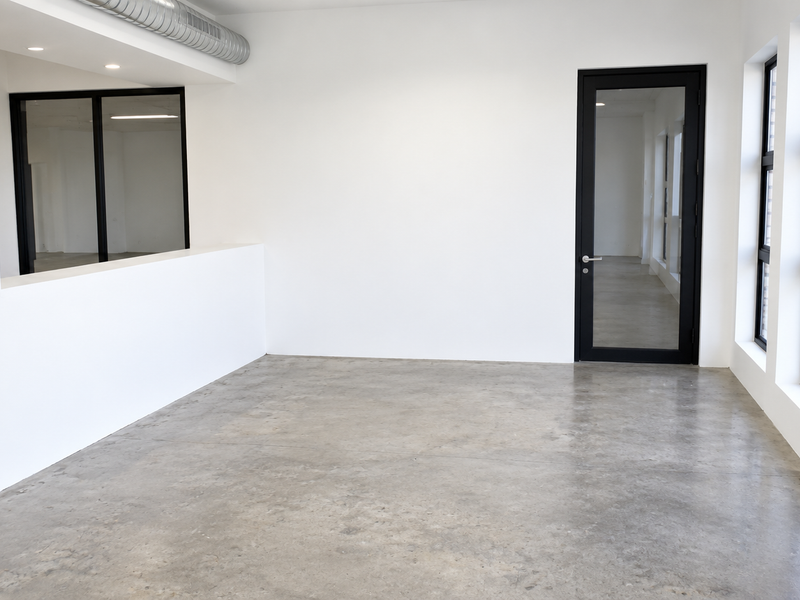 Modern, minimalist office space featuring polished concrete floors, exposed industrial ducting, and large black-framed windows providing ample natural light.