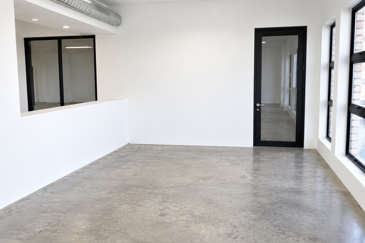 Modern, minimalist office space featuring polished concrete floors, exposed industrial ducting, and large black-framed windows providing ample natural light.