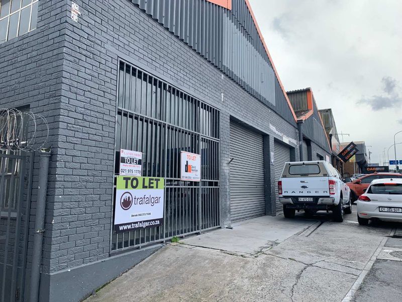 Front elevation of a commercial retail or industrial property featuring brick facade, metal roller shutter doors, and secure gated access.