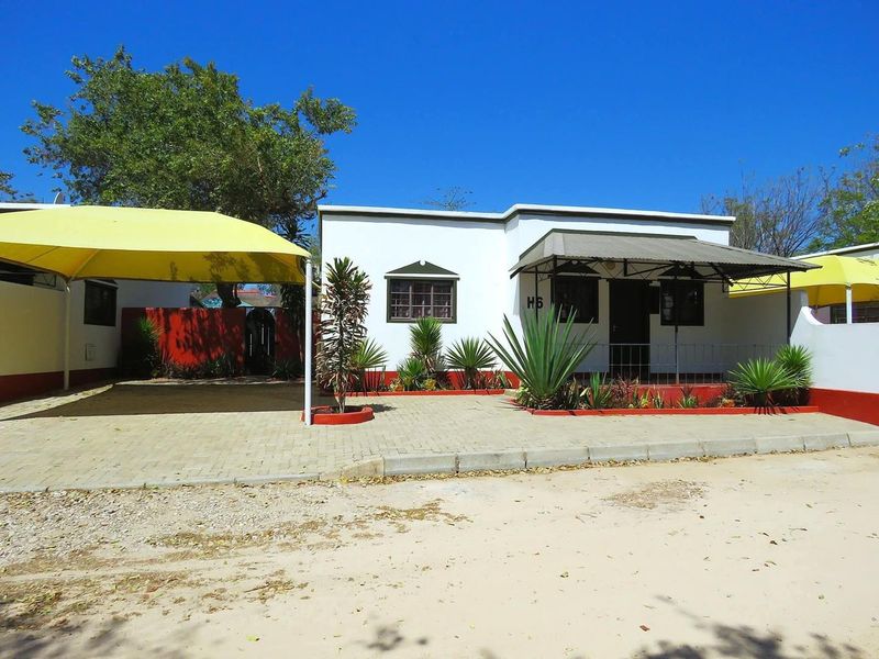 Front exterior view of a lodge unit featuring a paved driveway, shade sail carport, and a covered entrance porch with established garden beds.