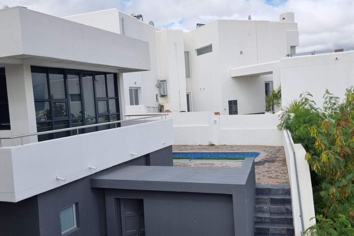 Modern multi-level residential exterior featuring a balcony, paved patio area, and a swimming pool set within a contemporary architectural design.