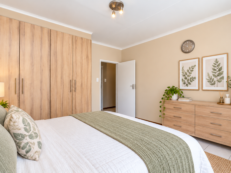 A well-maintained bedroom featuring light wood-finish built-in cupboards, neutral wall tones, and a functional layout with direct access to the hallway.