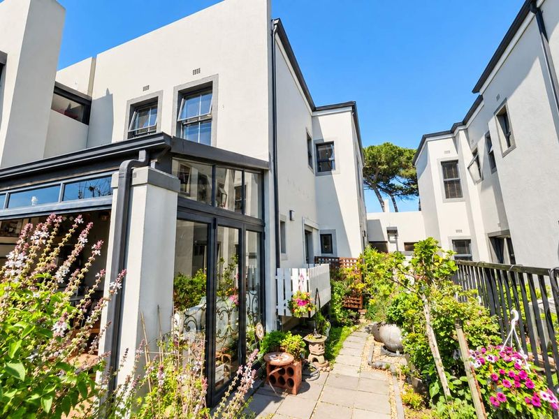 Rear view of a multi-story townhouse featuring a glass-enclosed patio, paved garden walkway, and secure metal perimeter fencing.