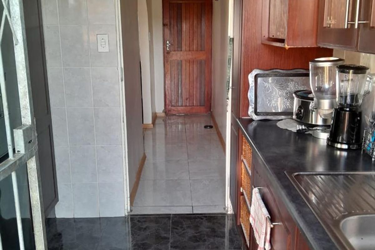 A functional kitchen featuring dark wood cabinetry, laminate countertops, and a stainless steel sink, leading into a tiled hallway.