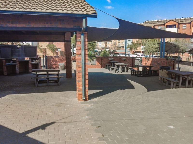 Outdoor entertainment area featuring a covered patio with a built-in braai, brick pillars, and shade sail coverage over a paved communal space.