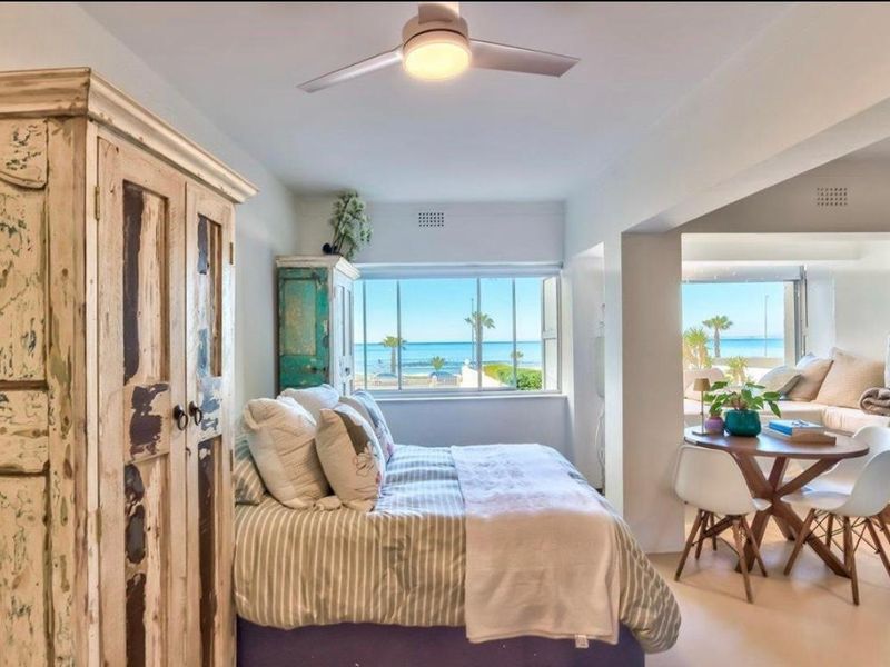 A bright bedroom featuring a ceiling fan and large windows with direct ocean views, creating a serene and airy coastal living space.