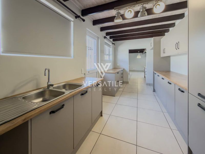 A clean, modern kitchen featuring light grey cabinetry, wooden countertops, double stainless steel sink, and exposed ceiling beams with track lighting.