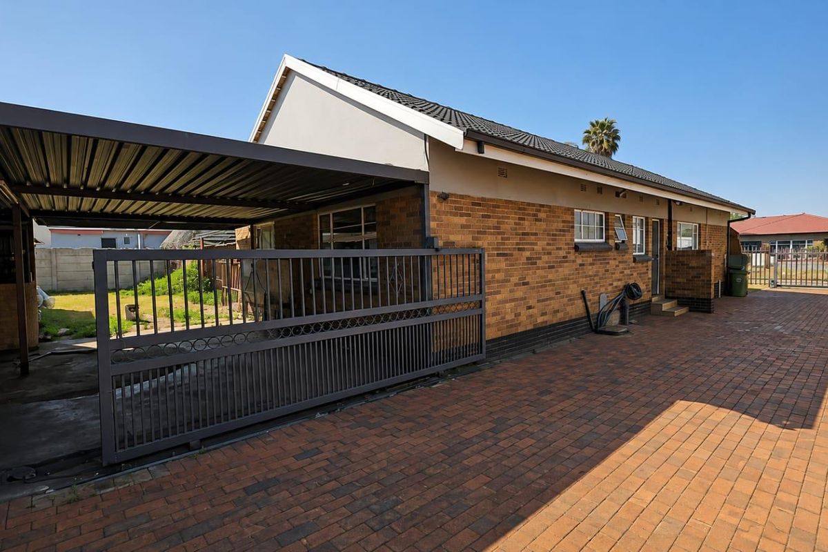 Rear exterior view of the house featuring a brick facade, a covered carport, a sliding security gate, and a paved driveway area.