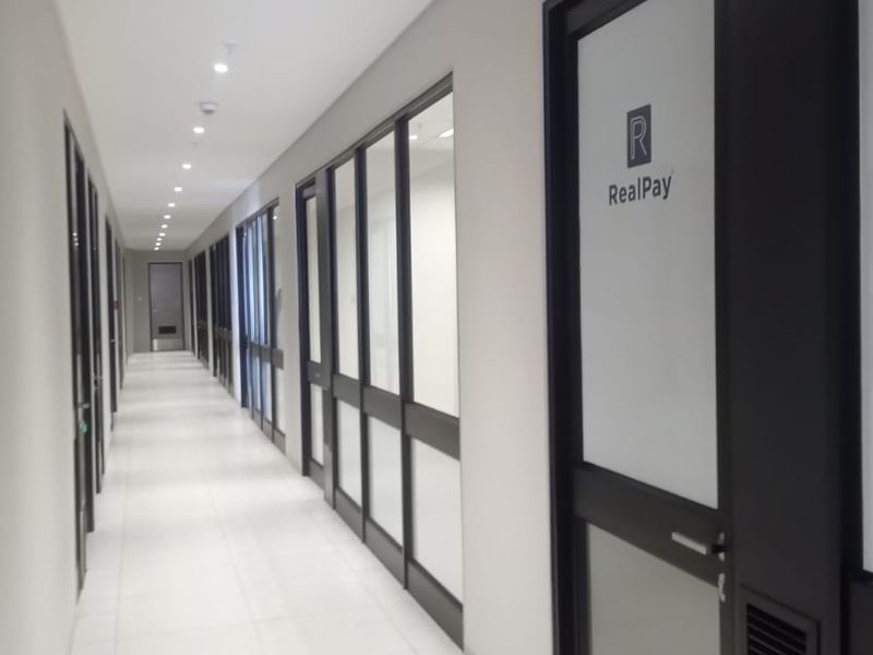 A modern, well-lit commercial office hallway featuring tiled floors, recessed lighting, and glass-partitioned office entrances with aluminium frames.