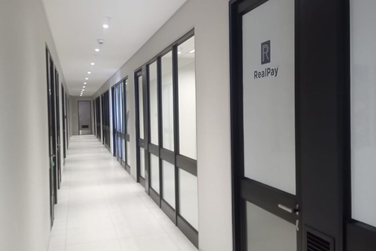 A modern, well-lit commercial office hallway featuring tiled floors, recessed lighting, and glass-partitioned office entrances with aluminium frames.