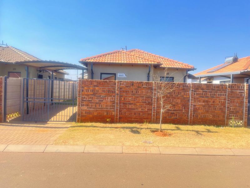 Front view of a residential property featuring a brick boundary wall, metal gate, paved driveway, and a carport under a tiled roof.