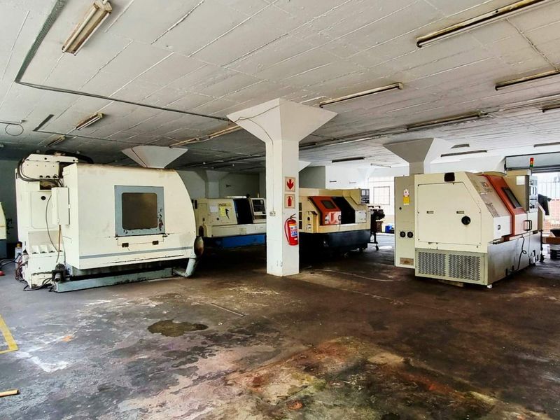 Industrial workshop space featuring concrete flooring, structural columns, and overhead fluorescent lighting, currently utilized for heavy machinery placement.