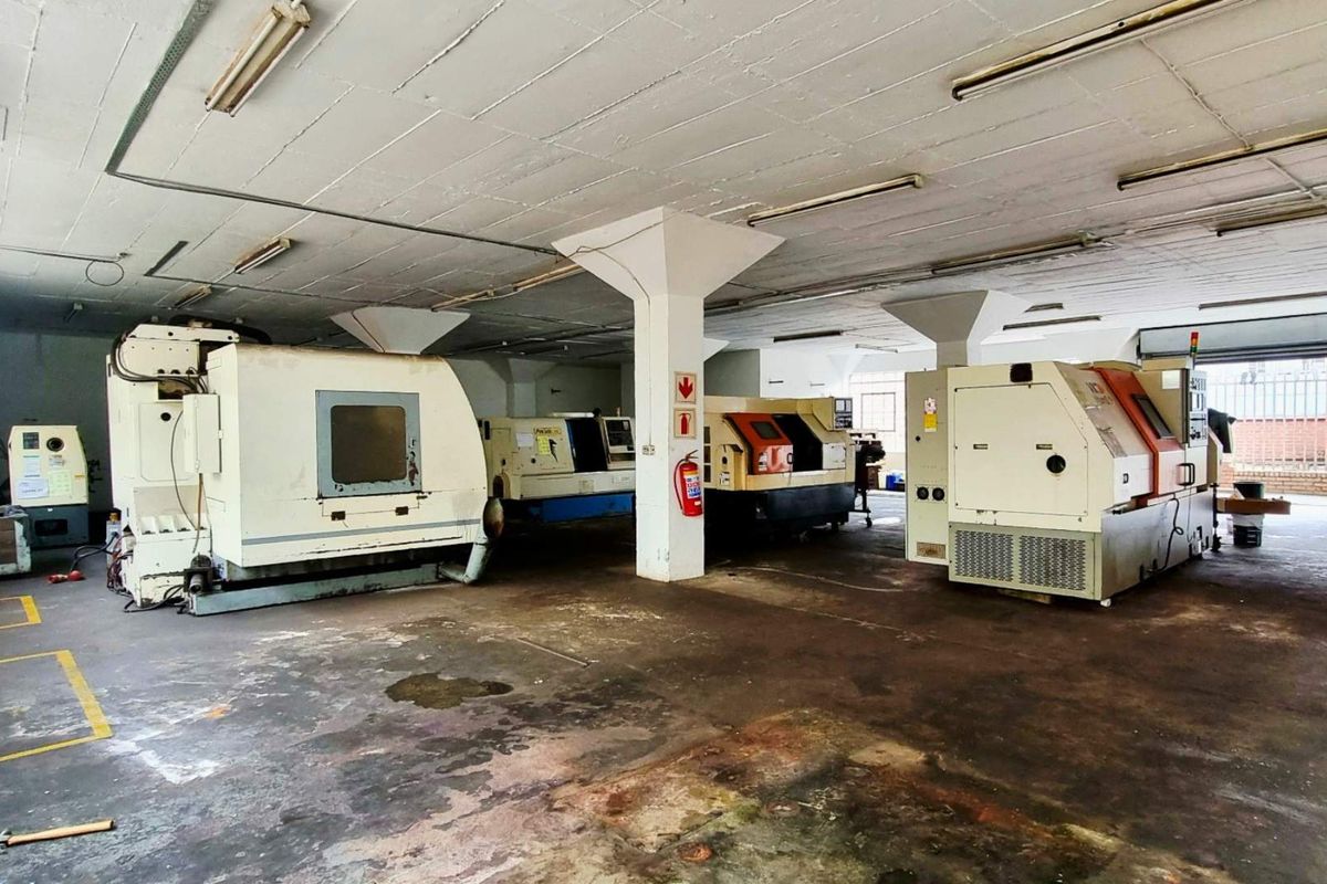 Industrial workshop space featuring concrete flooring, structural columns, and overhead fluorescent lighting, currently utilized for heavy machinery placement.